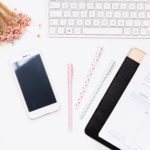 desktop with keyboard, pins, cell phone, and notebook.